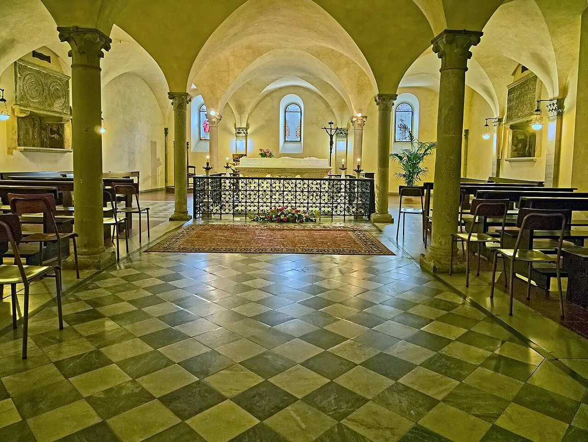 The vaulted crypt of Bobbio Abbey, the white sarcophagus of Saint Columbanus behind a wrought iron screen, early medieval carved slabs on the walls