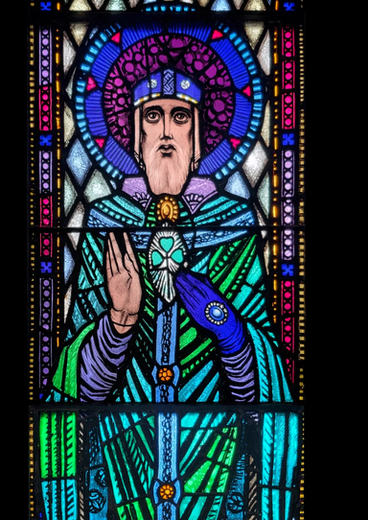 Harry Clarke's Saint Patrick in stained glass, a haloed bishop in blue and green vestments raising his hand and holding a white shamrock